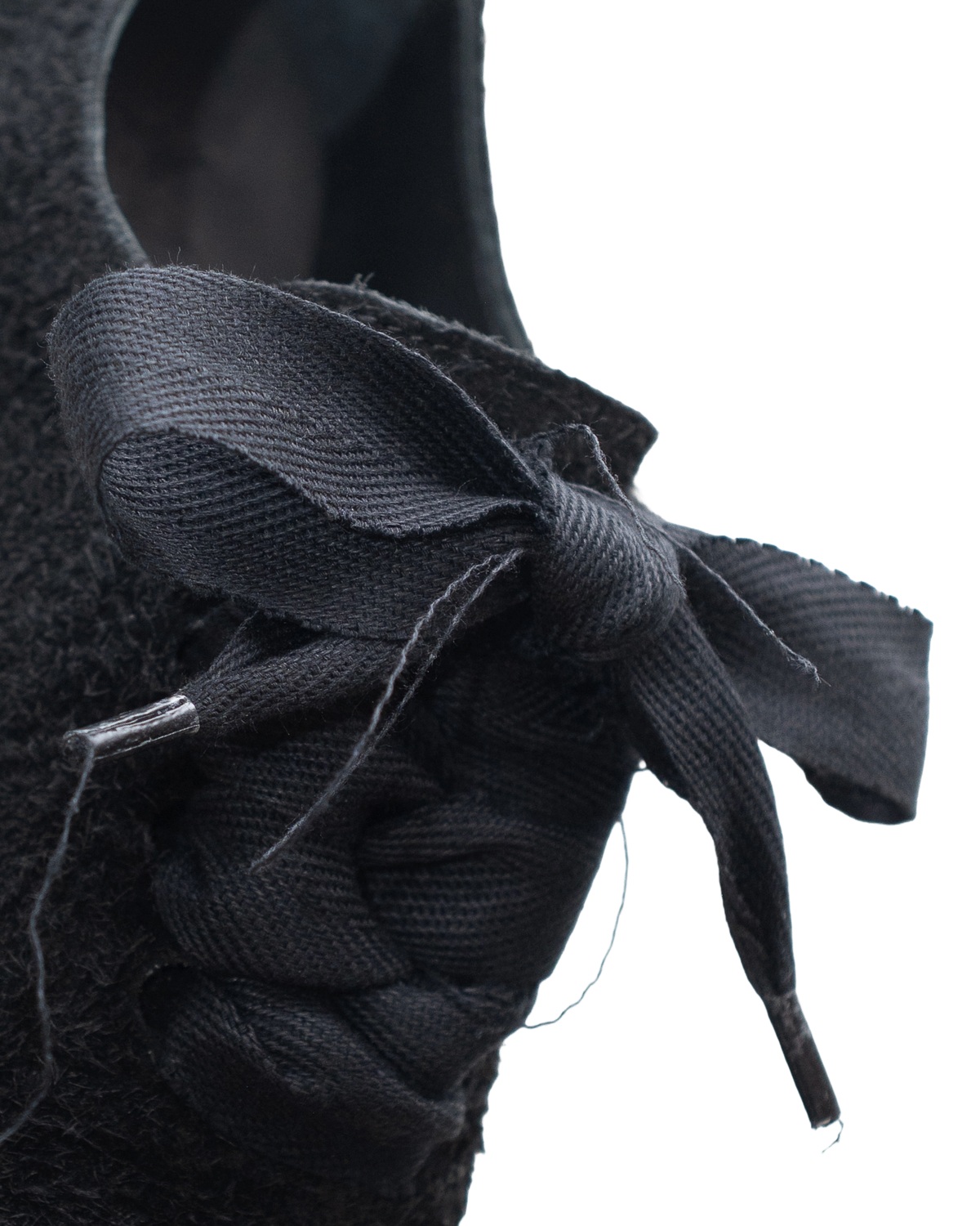Close-up of a black textured shoe featuring a braided lace-up detail and a large fabric bow on a plain white background.