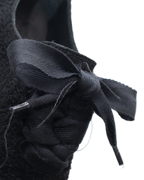 Close-up of a black textured shoe featuring a braided lace-up detail and a large fabric bow on a plain white background.